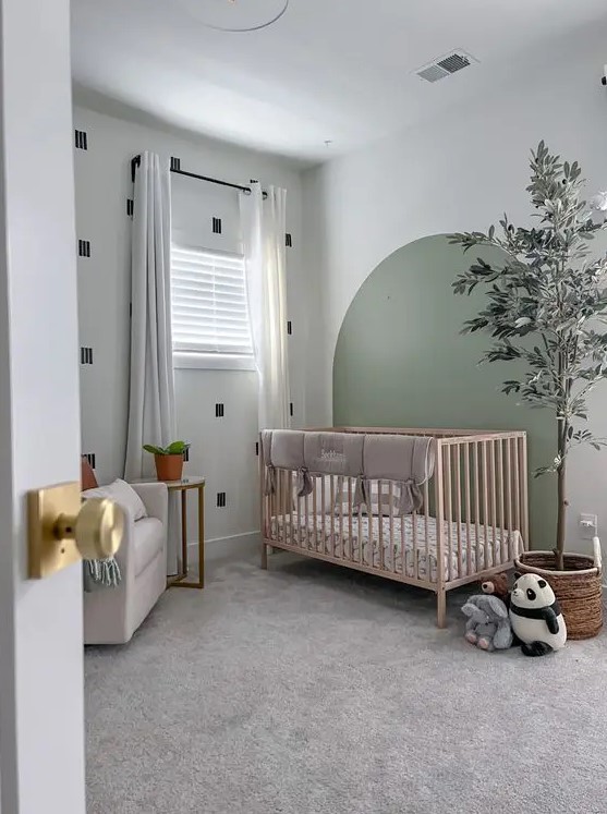 a small nursery with a printed wall and a sage green accent, a stained crib with neutral bedding, a potted tree and some toys