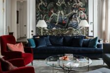 a sophisticated living room with a wall mural, a navy velvet sofa and red chairs, a round coffee table and a gorgeous chandelier