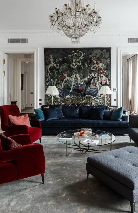 a sophisticated living room with a wall mural, a navy velvet sofa and red chairs, a round coffee table and a gorgeous chandelier