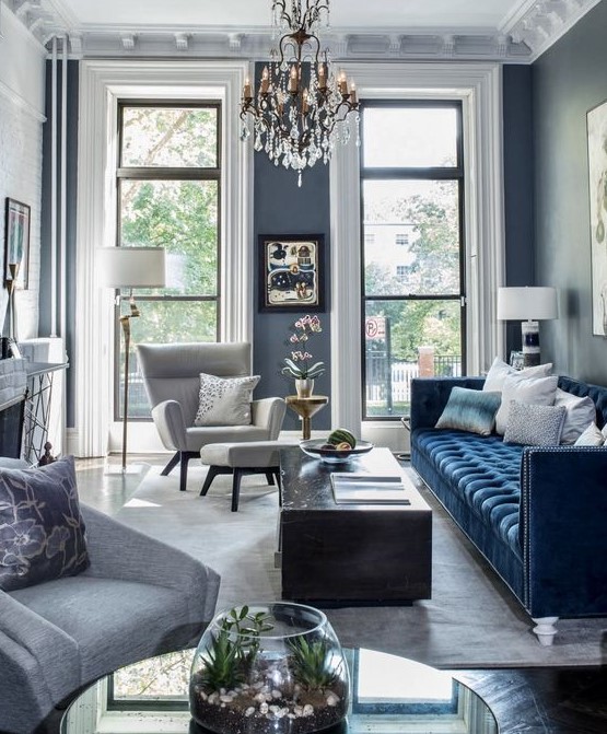 a sophisticated modern living room with grey walls, a navy sofa, a coffee table, a creamy chair with a footrest and a chic chandelier