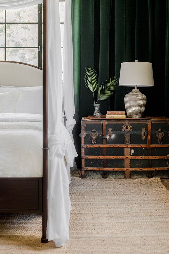 a sophisticated tropical bedroom with a framed bed with neutral bedding, a black vintage trunk as a nightstand, dark green curtains