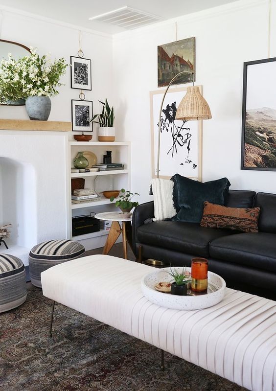 a stylish and catchy living room with a non-working fireplace, a black leather sofa, an upholstered bench, some shelves and artwork