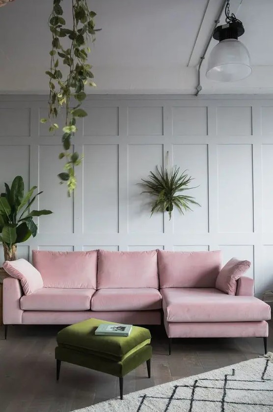 a stylish living room with dove grey paneled walls, a pink sectional sofa, a green pouf and some potted greenery around