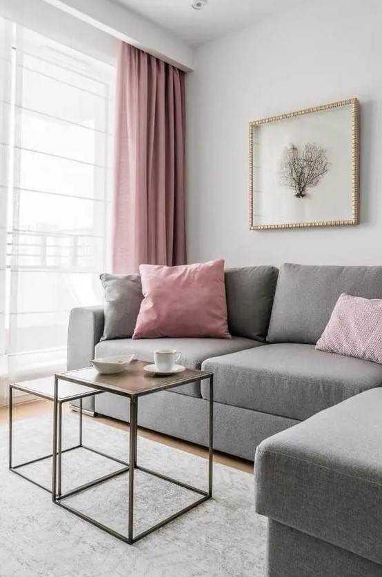 a very laconic living room with a grey sectional, pink and grey sofas, a couple of coffee tables and pink curtains