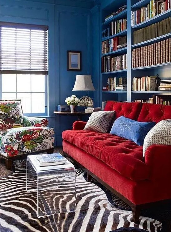 a vibrant space with blue walls, built-in bookshelves, a red sofa and a floral chair, layered rugs and an acrylic table