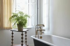 a vintage bathroom with a penny tile floor, a white casement window, a black clawfoot tub, a side table with a potted plant