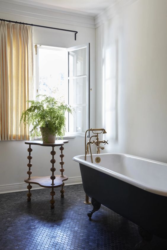 a vintage bathroom with a penny tile floor, a white casement window, a black clawfoot tub, a side table with a potted plant