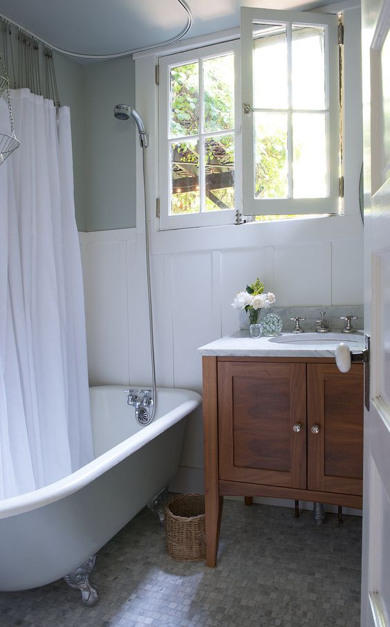 a vintage cottage bathroom with a casement window,  a grey clawfoot tub with a curtain, a stained console table and a basket