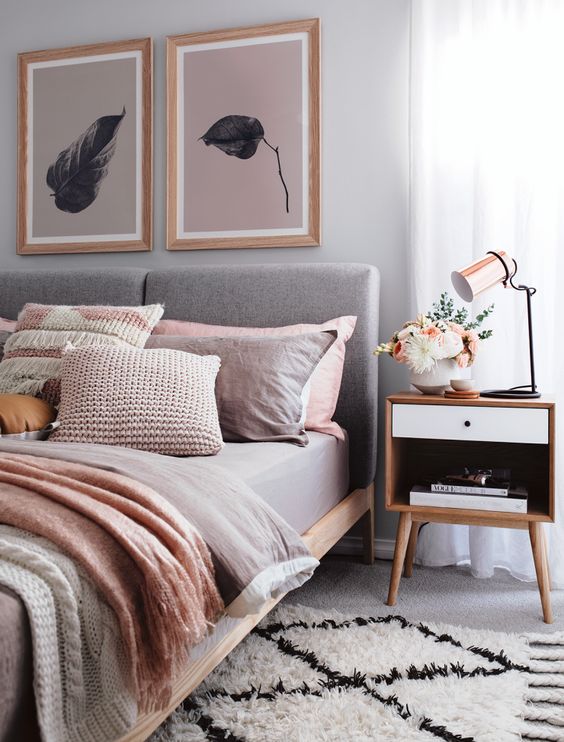 a welcoming bedroom with a grey upholstered bed and grey and pink bedding, a gallery wall, a nightstand with a rose gold lamp