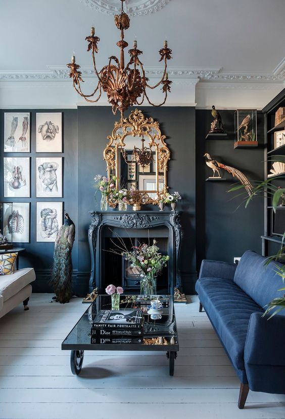 a whimsical living room with black walls, a vintage fireplace, a navy and a creamy sofa, an ornated frame mirror and a chandelier