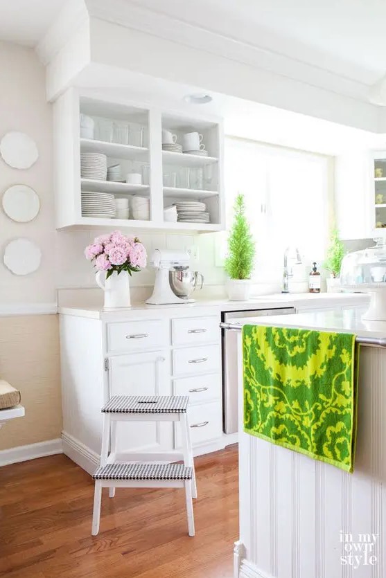 a white farmhouse kitchen with a large kitchen island, open upper cabinets, potted plants, an IKEA stool and bright linens