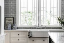 a white farmhouse kitchen with shaker cabinets, white stone countertops, a grey subway tile wall and a black kitchen island plus two casement windows
