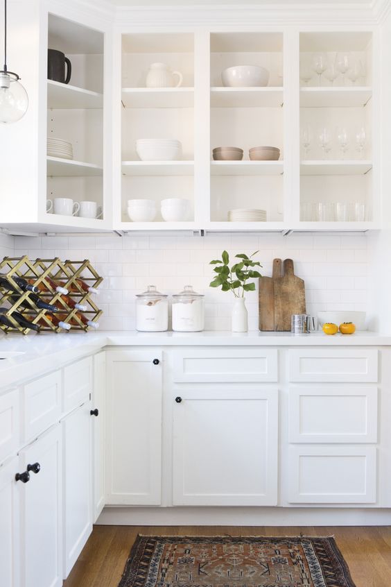 a white kitchen with open and shaker cabinets, white stone countertops and a white tile backsplash plus a brass wine bottle stand