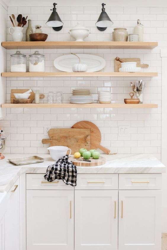 a white kitchen with shaker cabinets, white stone countertops, white subway tiles and open shelving instead of upper cabinets plus black sconces