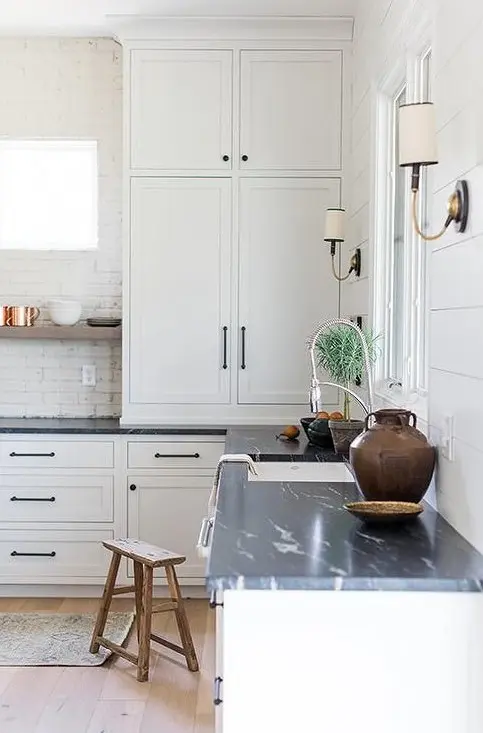 a white kitchen with shaker style cabinets, a brick backsplash, black soapstone countertops, black fixtures