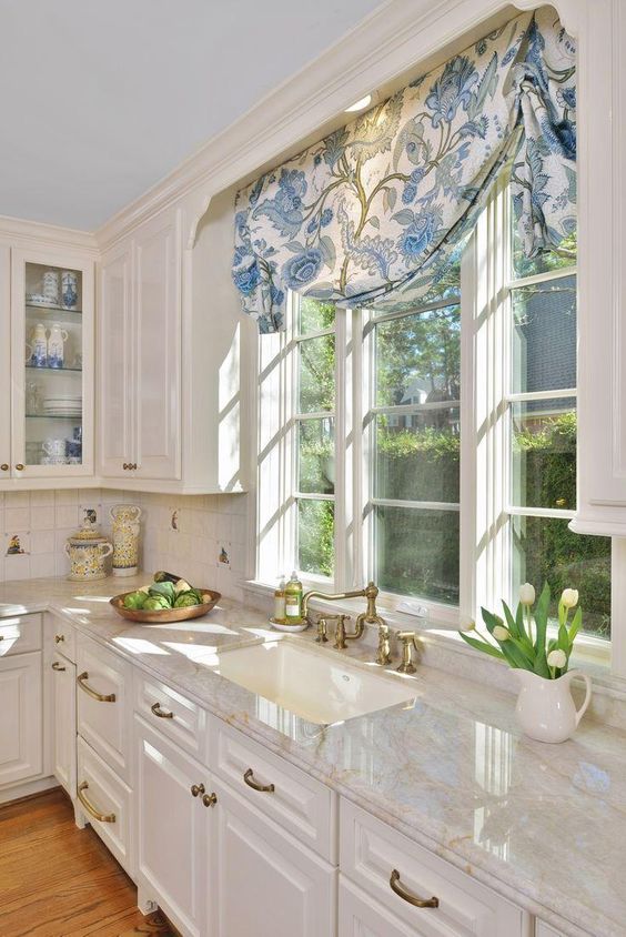 a white vintage kitchen with shaker cabinets, stone countertops, casement windows and printed curtains