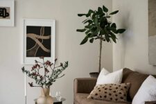 an airy living room with tan walls, a brown sofa, a white coffee table, abstract artworks, a potted tree and a pendant lamp
