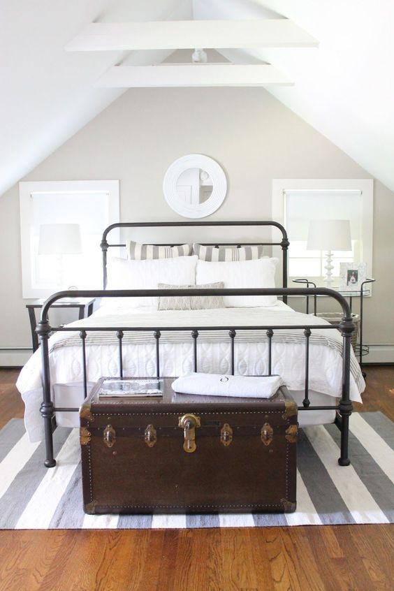 an attic guest bedroom with a black metal bed, neutral bedding, a dark-stained vintage chest and a striped rug
