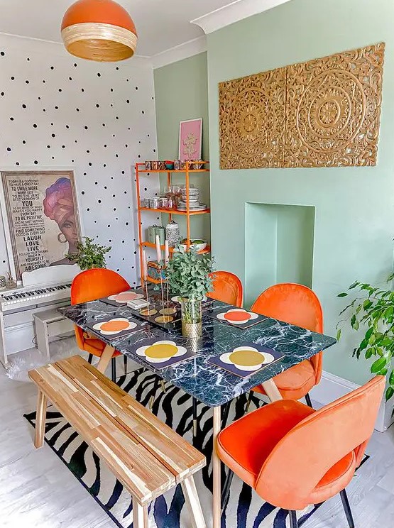 an eclectic boho dining room with a mint green accent wall, an orange shelving unit, orange chairs, a dark table and a bench
