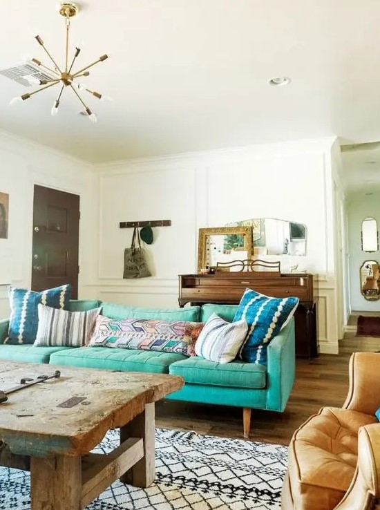 an eclectic living room with a mid-century modern turquoise sofa, a rough wooden table, a chair, a wooden dresser and some mirrors on it