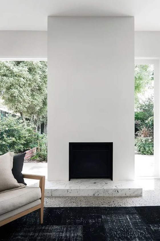 an ultra minimalist space with a white fireplace with a marble detail, a black rug and a neutral chair plus a garden view