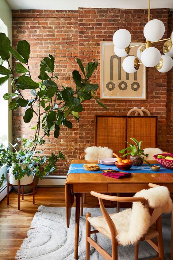 a mid-century modern meets boho dining room with a brick accent wall, a stained table and chairs, a rattan storage unit, potted plants and a chandelier