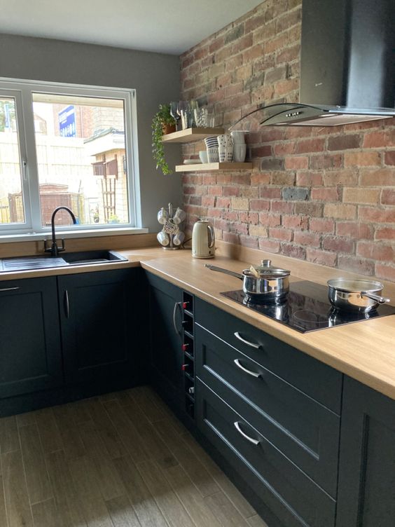 a modern black kitchen with butcherblock countertops, a red brick accent wall, black appliances and black fixtures and a stainless steel hood
