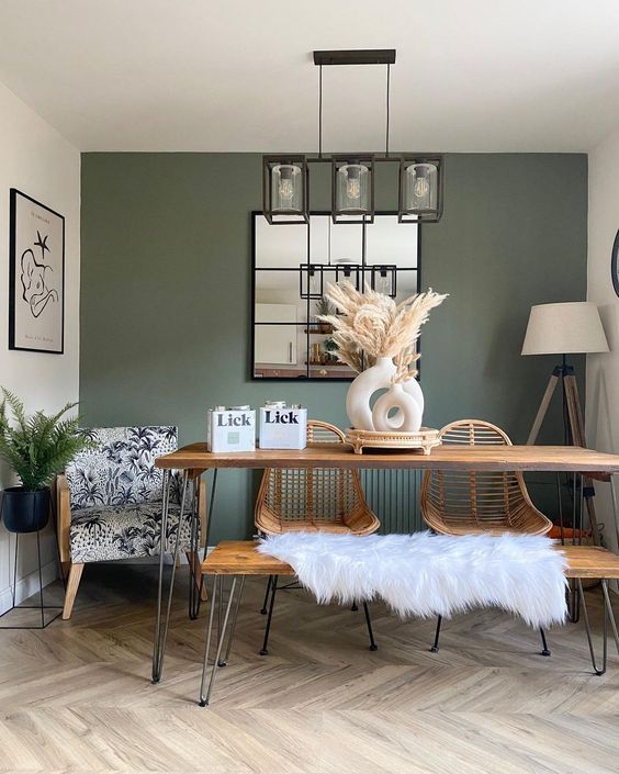 a cool boho mid-century modern dining space with a green accent wall, a hairpin leg table and a bench, a rattan chairs, a candelier and a mirror