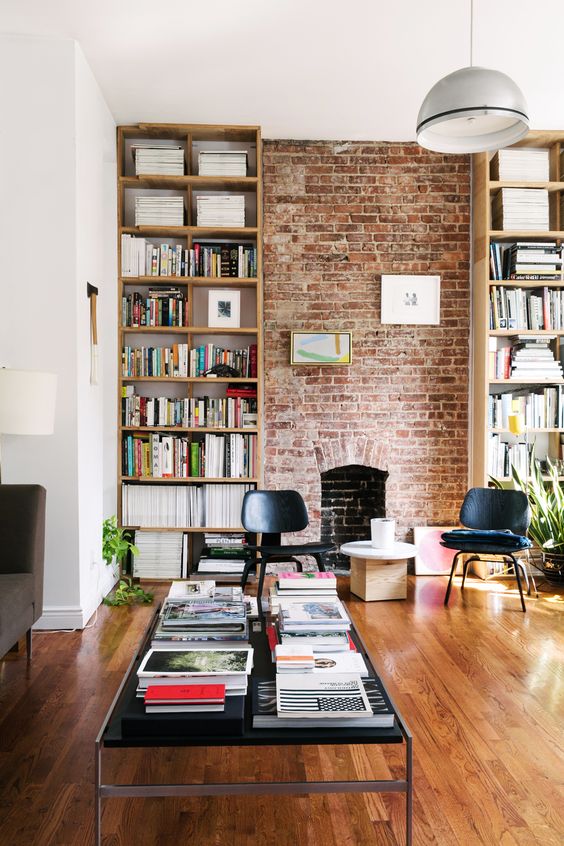a living room with a red brick wall, tall bookcases, a black coffee table, black chairs, potted plants and a pendant lamp