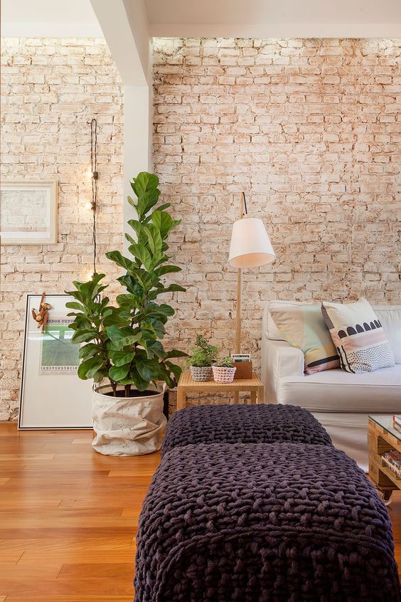 a living room with a whitewashed brick wall, a neutral sofa and grey poufs, side and coffee tables, potted plants and lamps
