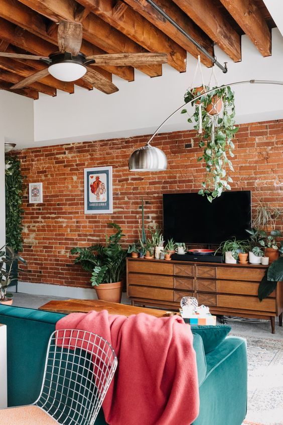 a mid-century modern living room with a brick accent wall, a stained TV unit, a green sofa and a coral blanket, potted plants
