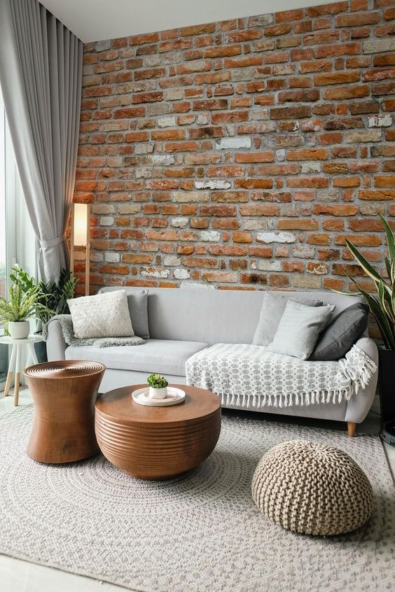 a neutral living room with a brick accent wall, a grey sofa and pillows, coffee tables and a knit pouf, dove grey curtains
