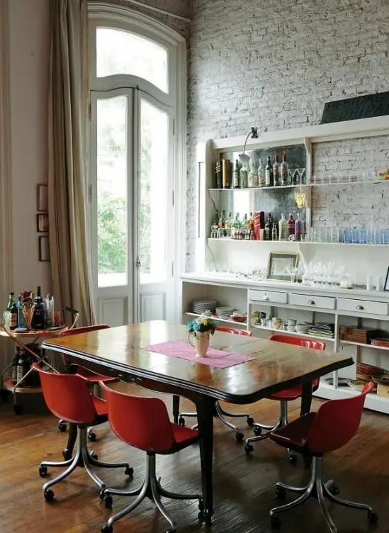 an eclectic dining space with a whitewashed brick wall, a large buffet and shelves, a refined wooden table and modern red chairs