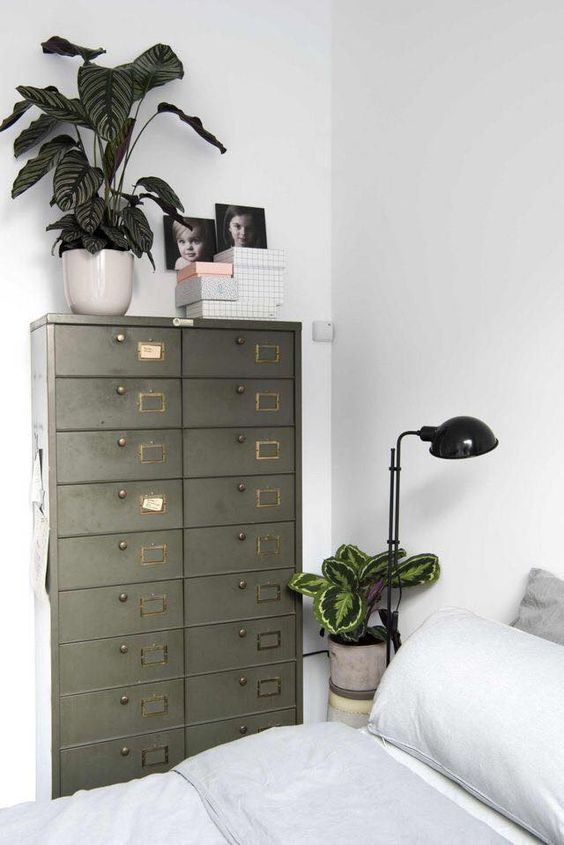 a Scandinavian bedroom with a bed with neutral bedding, a table lamp and some plants, a green card cabinet as a storage unit