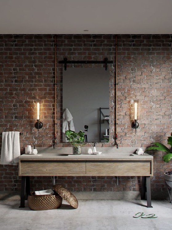 a red brick wall contrasts the sleek vanity, wall lamps and copper pipes isntead of a usual mirror frame