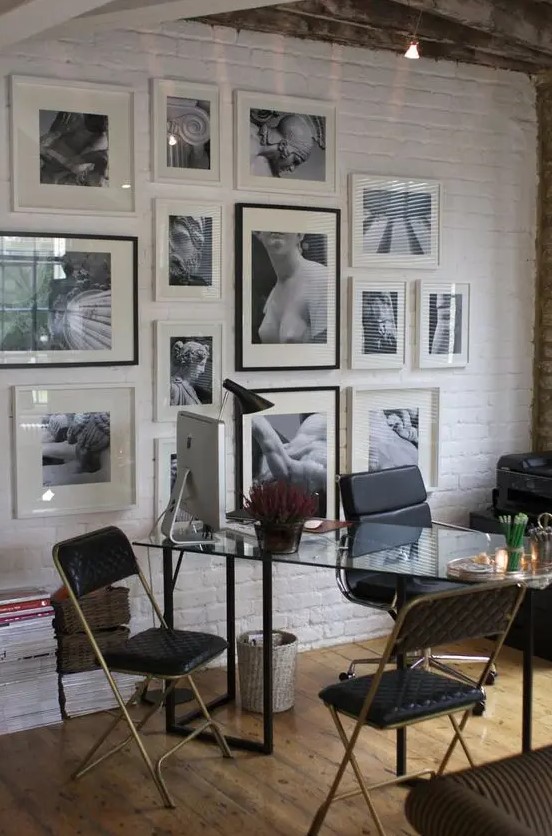 a home office with a white brick accent wall, a glass desk, black chairs, a black and white gallery wall and some lights