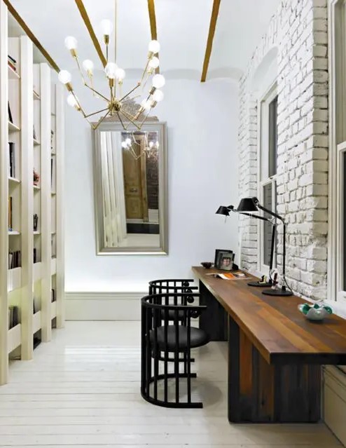 a neutral home office with a large shelving unit, a textural white brick wall, a large stained shared desk, black chairs, a mirror and a chandelier