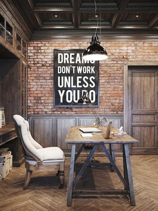 a rustic meets industrial home office with a red brick accent wall, dark-stained furniture, a trestle desk, a neutral chair and a black pendant lamp