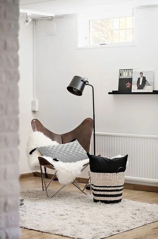 a Scandinavian nook with a brown leather butterfly chair, a blanket and a pillow, a basket with more pillows and a black floor lamp