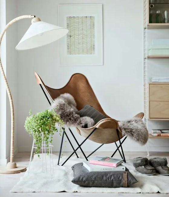 a Scandinavian nook with a brown leather butterfly chair, a neutral rug and a pillow, a floor lamp and a trellis with greenery