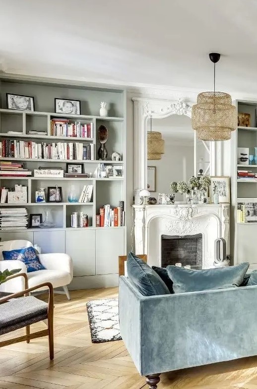 a beautiful and light-filled French chic living room with grey bookcases, a beautiful French fireplace, a slate blue sofa and some pretty modern chairs