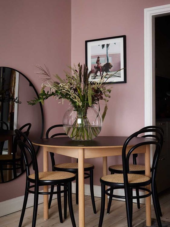 a beautiful dining space with mauve walls, a round mirror, a black and stained table and chairs, an artwork is cool