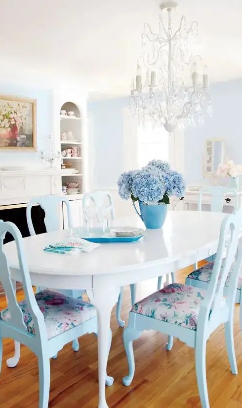 a beautiful pastel blue and white dining room with a pale blue accent wall, a fireplace, an oval table, blue chairs with floral upholstery and a crystal chandelier
