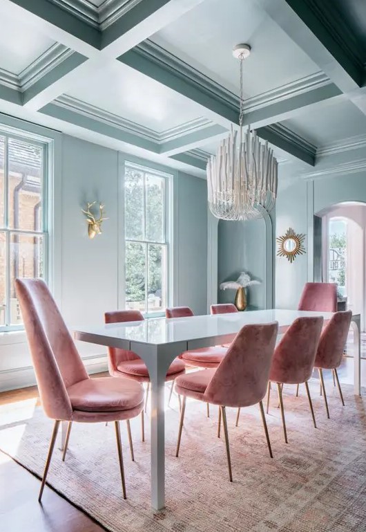 a beautiful pastel blue dining room with a coffered ceiling, large windows, a corner niche, a blue table and pink chairs