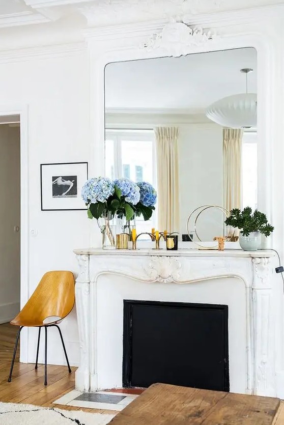 a beautiful white ornated Franch fireplace with an oversized mirror on top, a plywood chair and a wooden coffee table