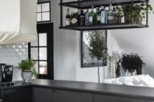 a black industrial kitchen with sleek cabinets, a suspended shelf over the kitchen island, black beams and greenery around