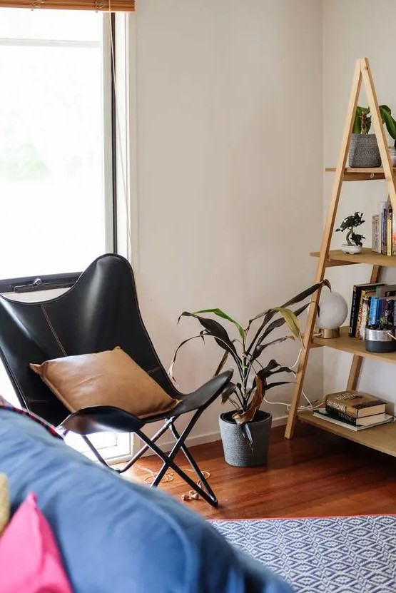 a black leather butterfly chair, a tan pillow, a ladder-style bookshelf and a printed rug for a mid-century modern space