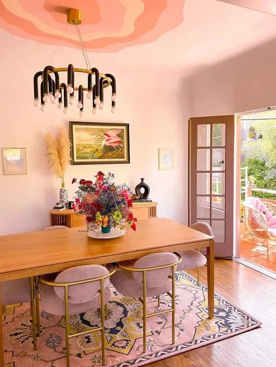 a blush dining space with a stained table and dusty pink chairs, a pink printed rug, a stained credenza, a black chandelier