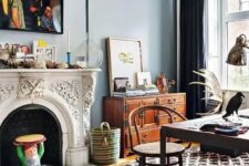 a boho eclectic home office with blue walls, an ornated French fireplace, printed layered rugs, a dark desk and a rattan chair, a stained sideboard