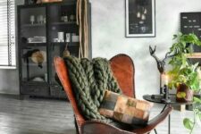 a bold and catchy space with a brown leather butterfly chair, a green chunky blanket, a black display unit and a side table with greenery